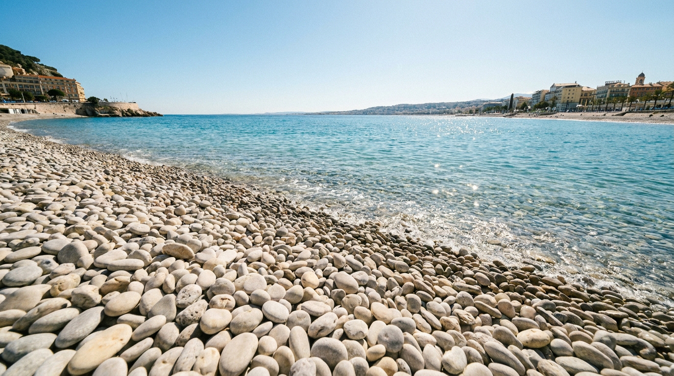 Plage de galets à Nice avec mer bleue scintillante, le long du littoral bordé de bâtiments sous un ciel clair.